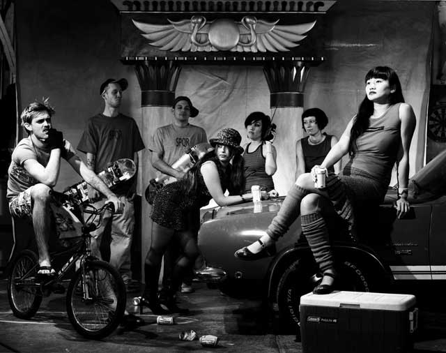 Hipters kicking it and drinking beer. Glamorous woman sits on the hood of an old Plymouth